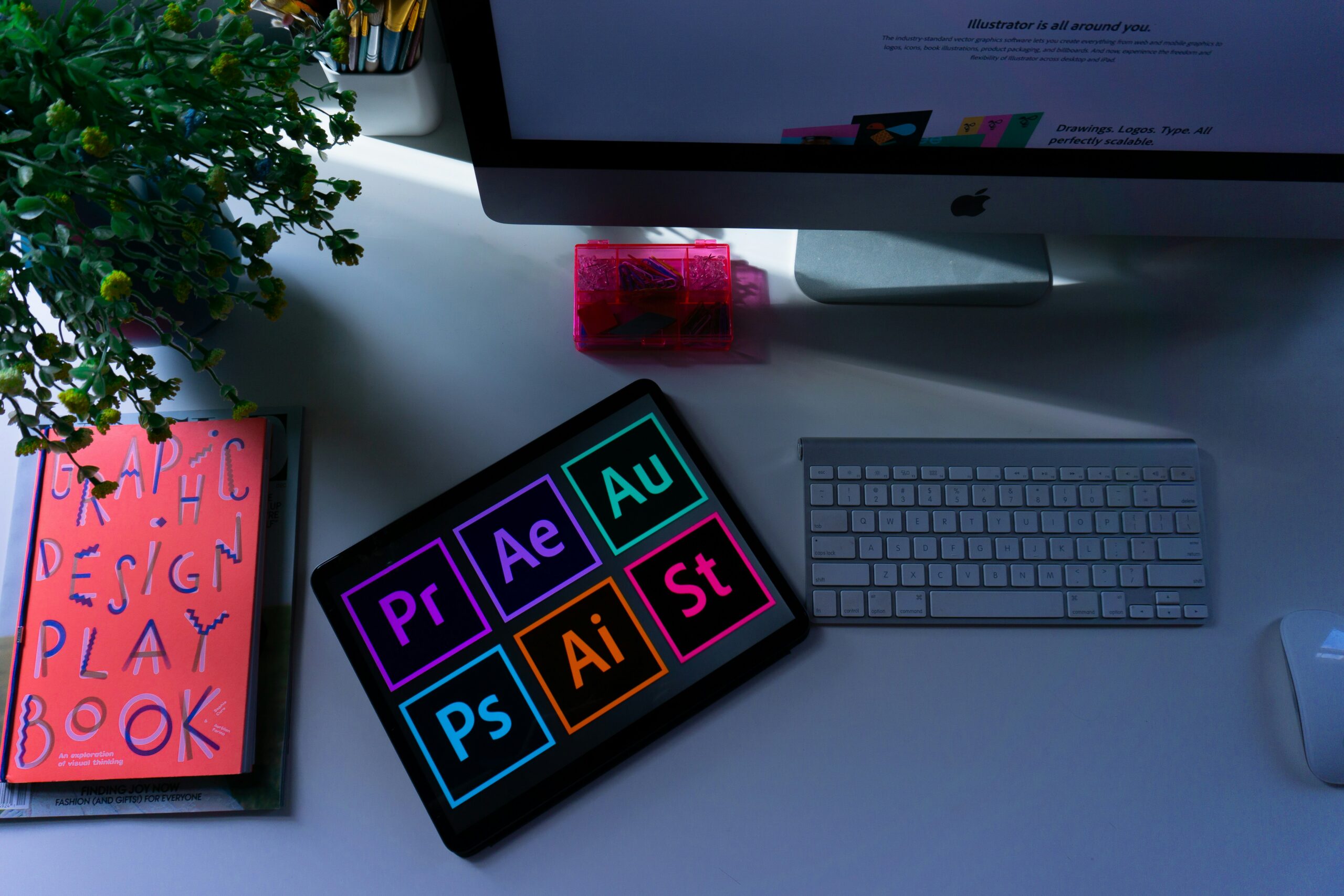 A workspace with a lit computer monitor, wireless keyboard, and graphic design tools. A plant and book add color, evoking creativity and productivity.