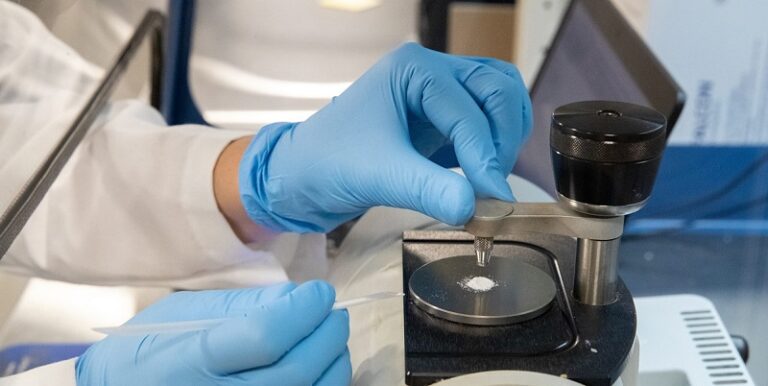 A scientist with blue gloves adds powder to a metallic plate on lab equipment, suggesting precision and focus in a laboratory environment.