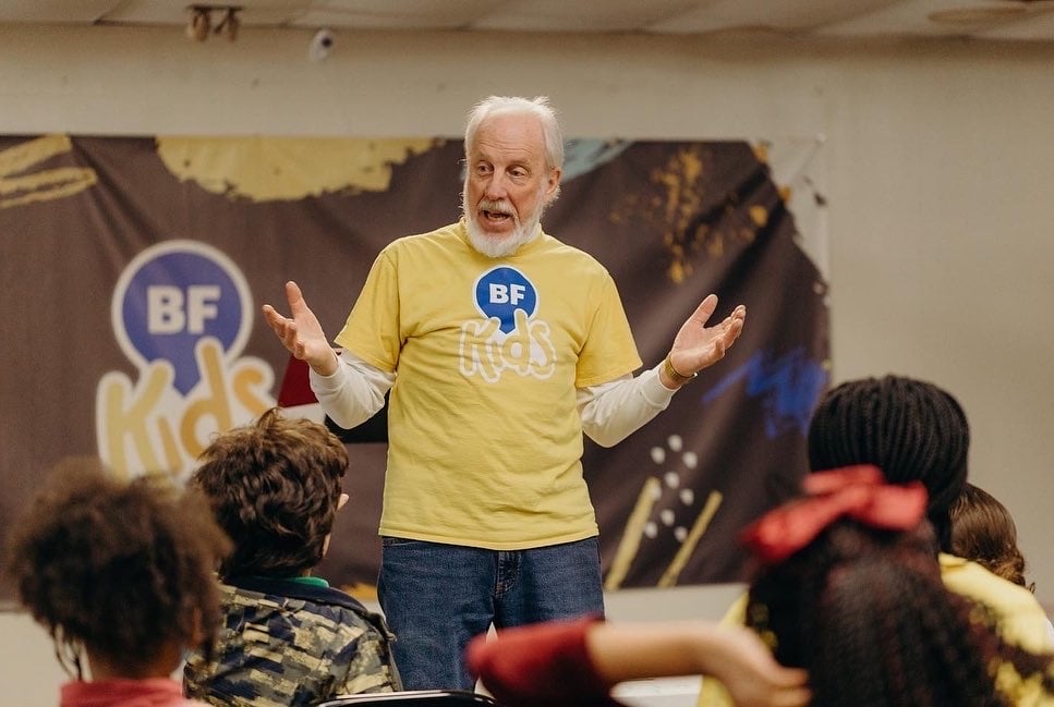 Elderly man in a yellow "BF Kids" shirt gestures engagingly while speaking to children in a classroom, fostering an educational and friendly atmosphere.