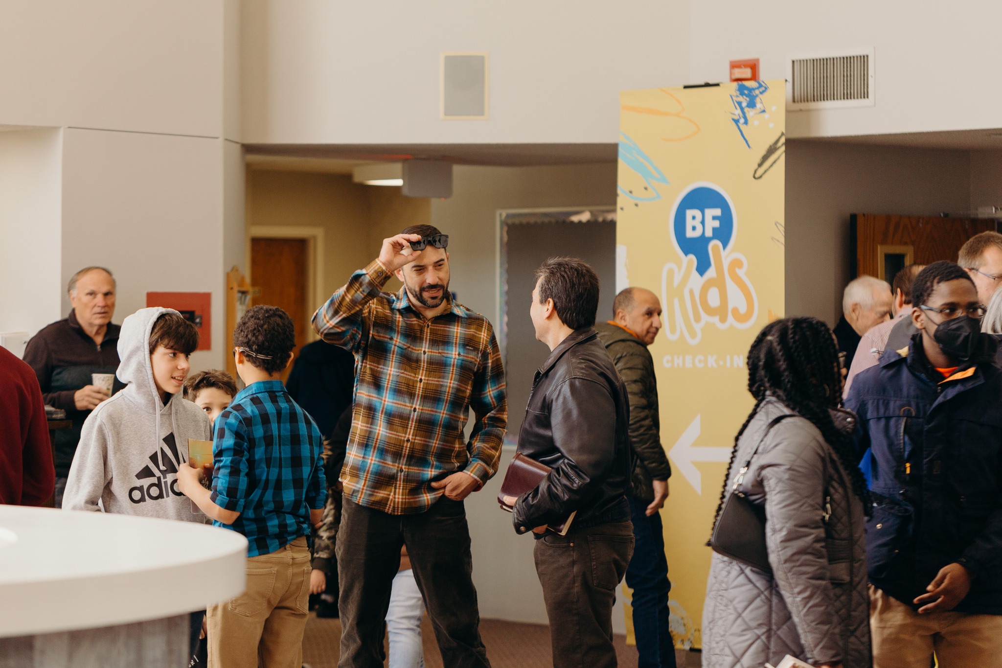 A group of people socialize in a lobby near a "BF Kids Check-In" sign. The mood is friendly and relaxed, with diverse individuals mingling.