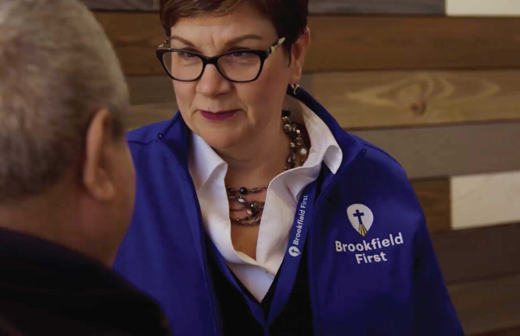 A woman wearing glasses and a blue "Brookfield First" jacket is engaged in conversation with a person facing away. The setting appears warm and conversational.