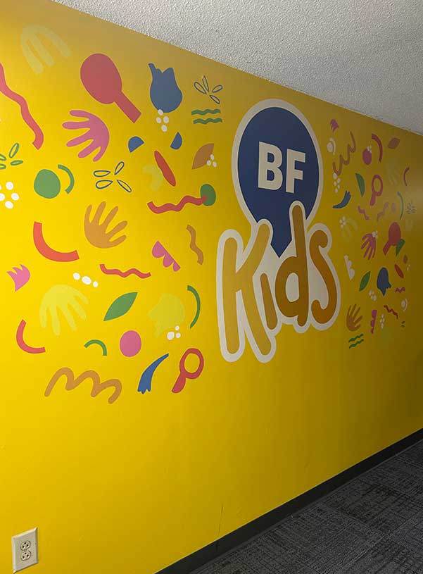 Bright yellow wall with colorful, playful shapes scattered around the words "BF Kids" in bold letters, conveying a cheerful and vibrant atmosphere.