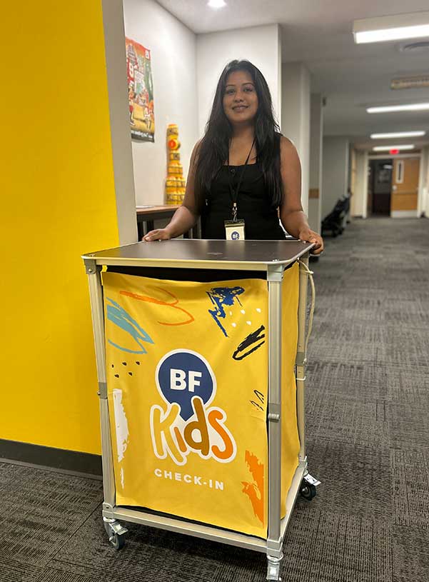 A smiling woman stands behind a yellow cart with "BF Kids Check-In" written on it in bold letters. The hallway has colorful decor, creating a welcoming atmosphere.
