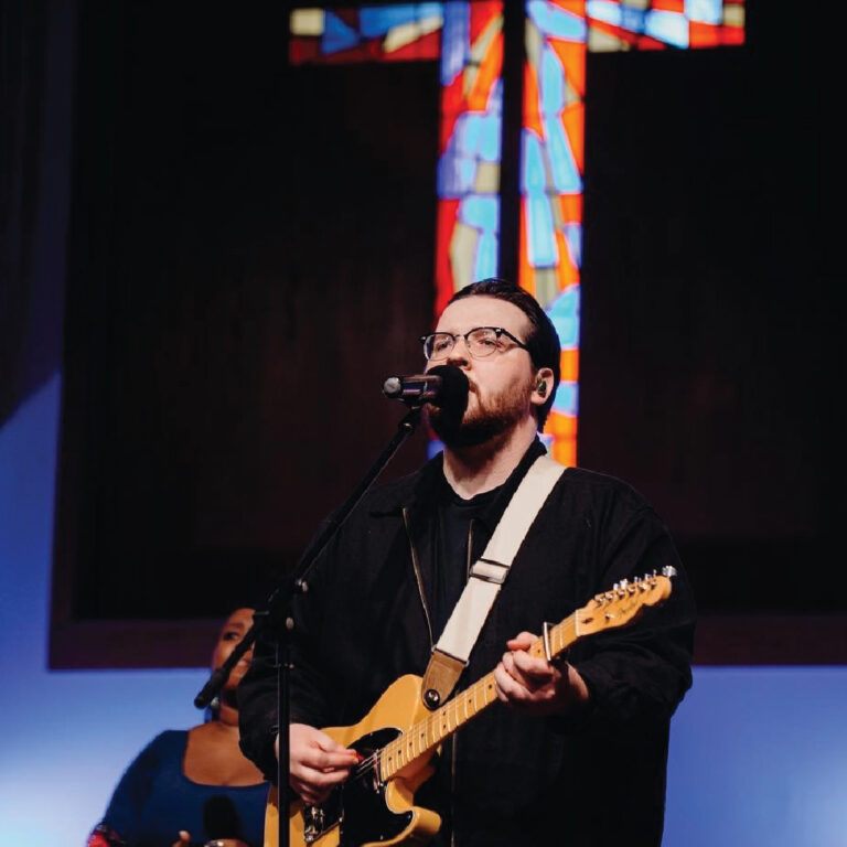 A musician with glasses plays an electric guitar and sings into a microphone. Behind is a colorful stained glass cross, conveying a serene and spiritual atmosphere during church worship.