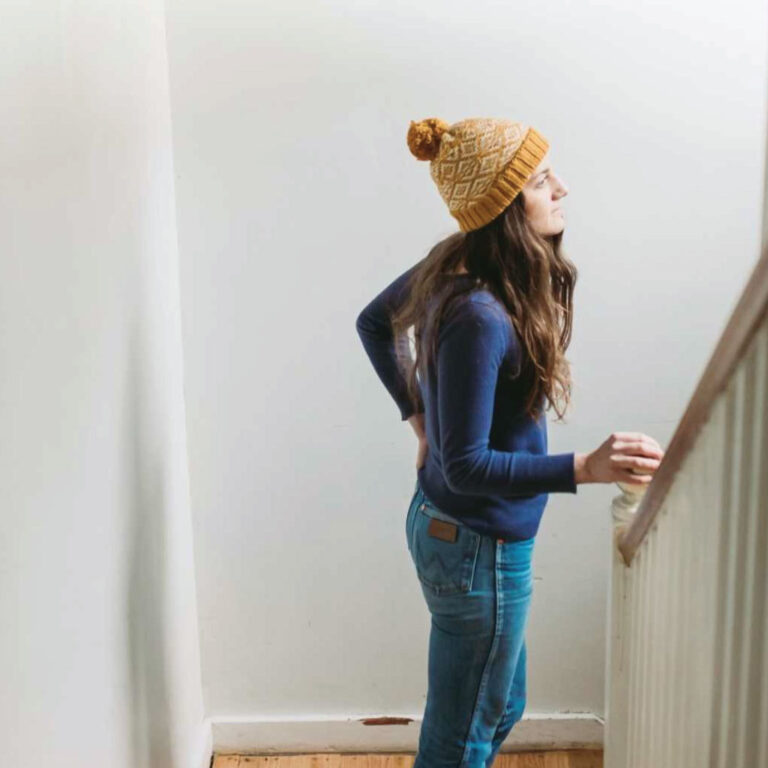 A woman in a mustard knit hat and blue sweater leans on a stair railing. She gazes upwards with a serene expression, in a softly lit room.