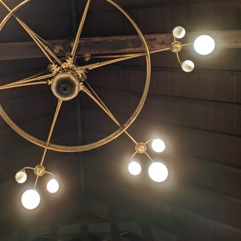 Antique chandelier with a circular metal frame and three arms, each holding glowing bulbs. The setting is dim, highlighting the chandelier’s ornate design.