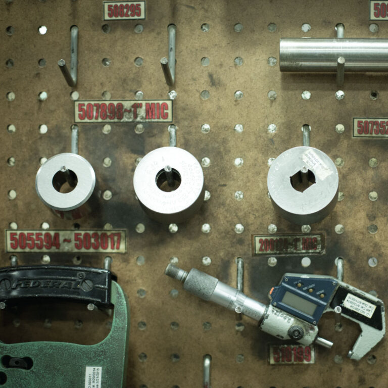 A brown pegboard holds metal rings and a digital caliper with labels. Industrial setting, precise tools hinting at accuracy and organization.